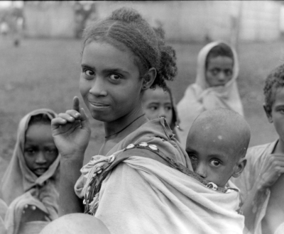 Woman looks into camera with baby in a sling on her back, and other children behind her also looking at the camera in this black and white photo