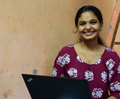 Previously sponsored girl by World Vision smiling and looking at the camera in her home in India while working on her laptop