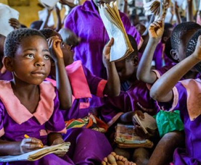 A girl from Malawi in a school uniform with a notebook on her lap listening to the class, with lots of classmates behind her