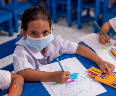 Girls from the Philippines wearing a face mask and school uniform looks up to the camera while drawing at school