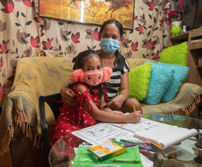 Girl from Philippines wearing a pink facemask sitting next to her mother at a table, also wearing a facemask.