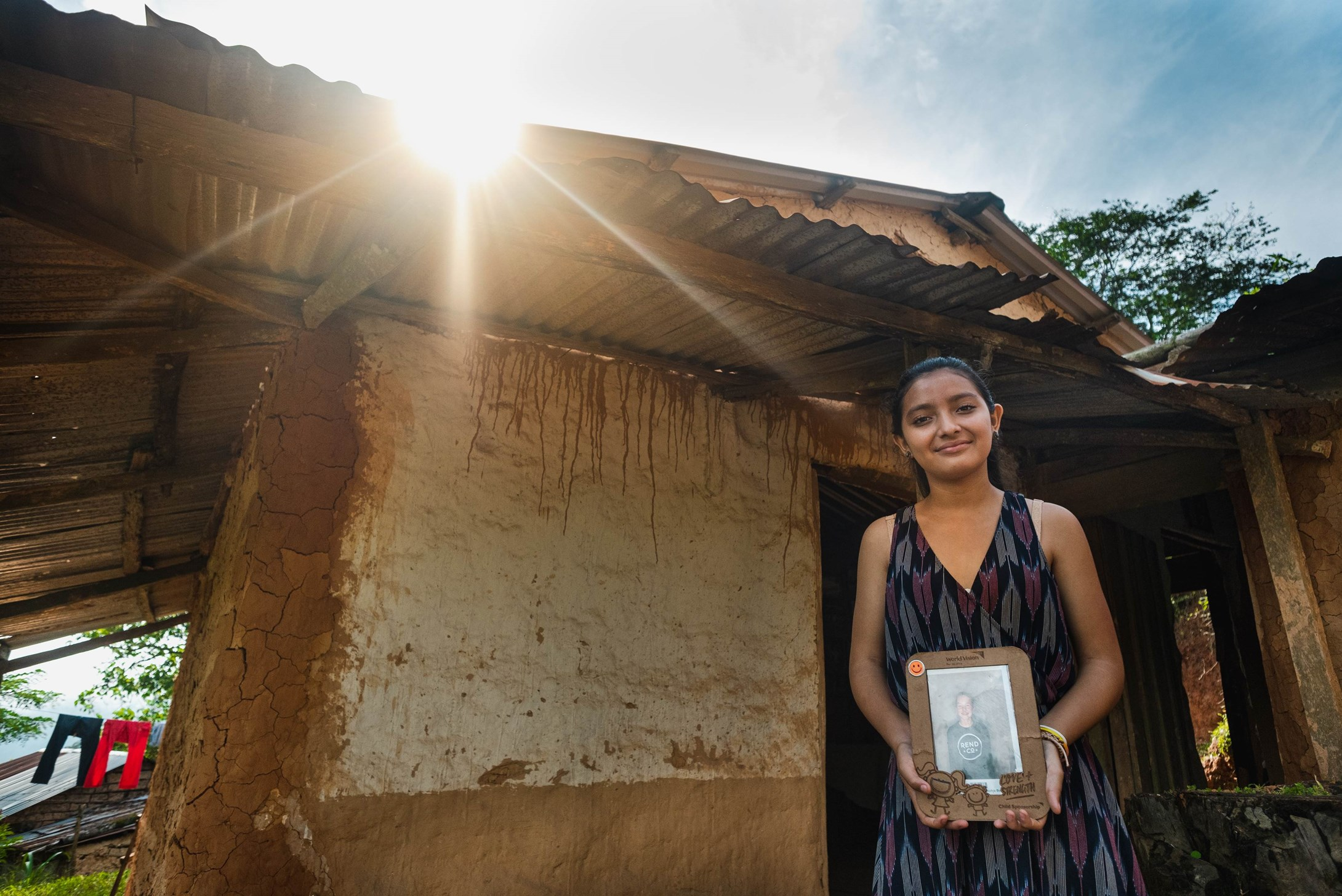 Sponsored girl from Honduras standing outside her house and holding up a portrait photo of her sponsor.