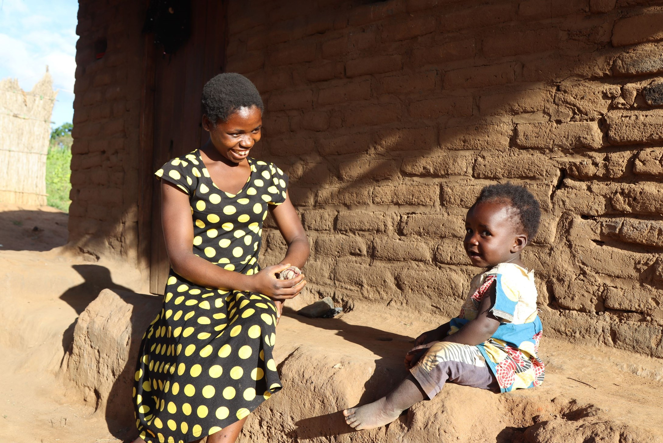 Teen mother from Malawi sitting on concrete outside her house and smiling to her 15-month old toddler