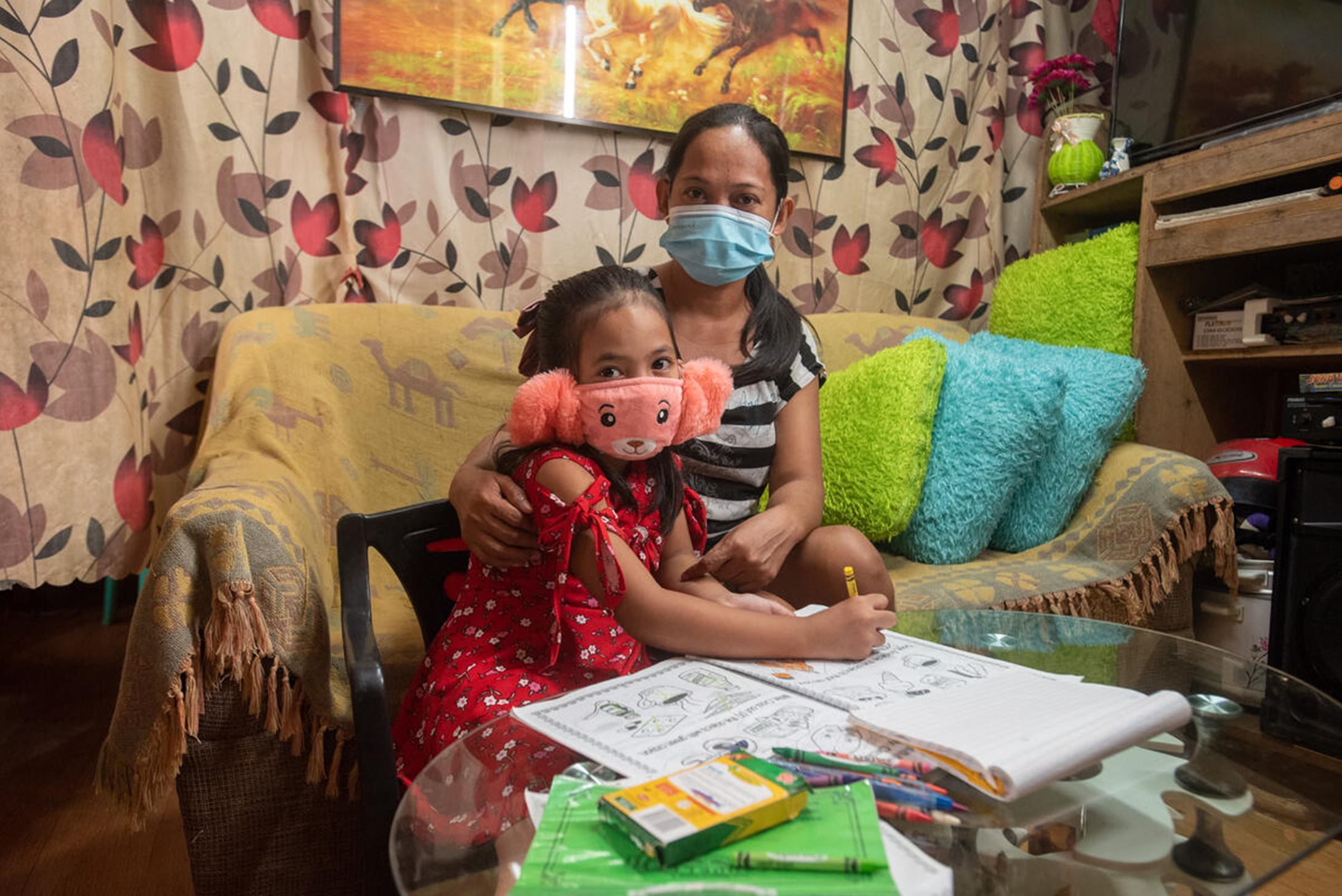 Girl from Philippines wearing a pink facemask sitting next to her mother at a table, also wearing a facemask.