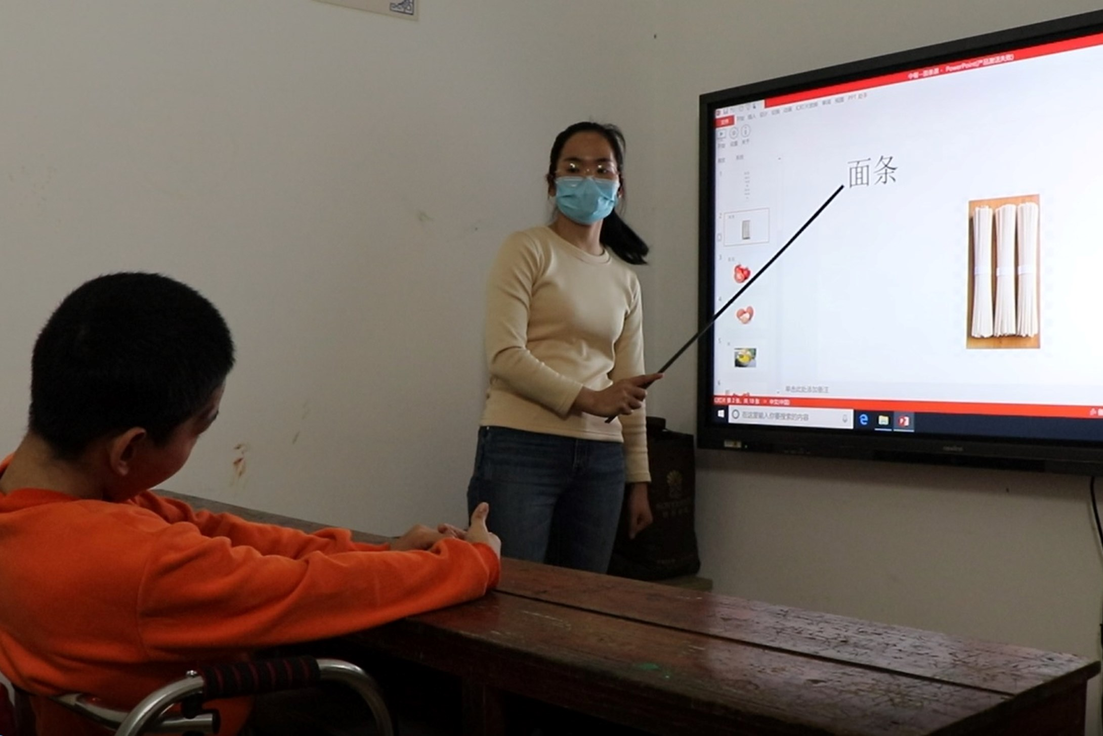 Woman wearing a facemask teaching a child on a projected whiteboard in a classroom in China