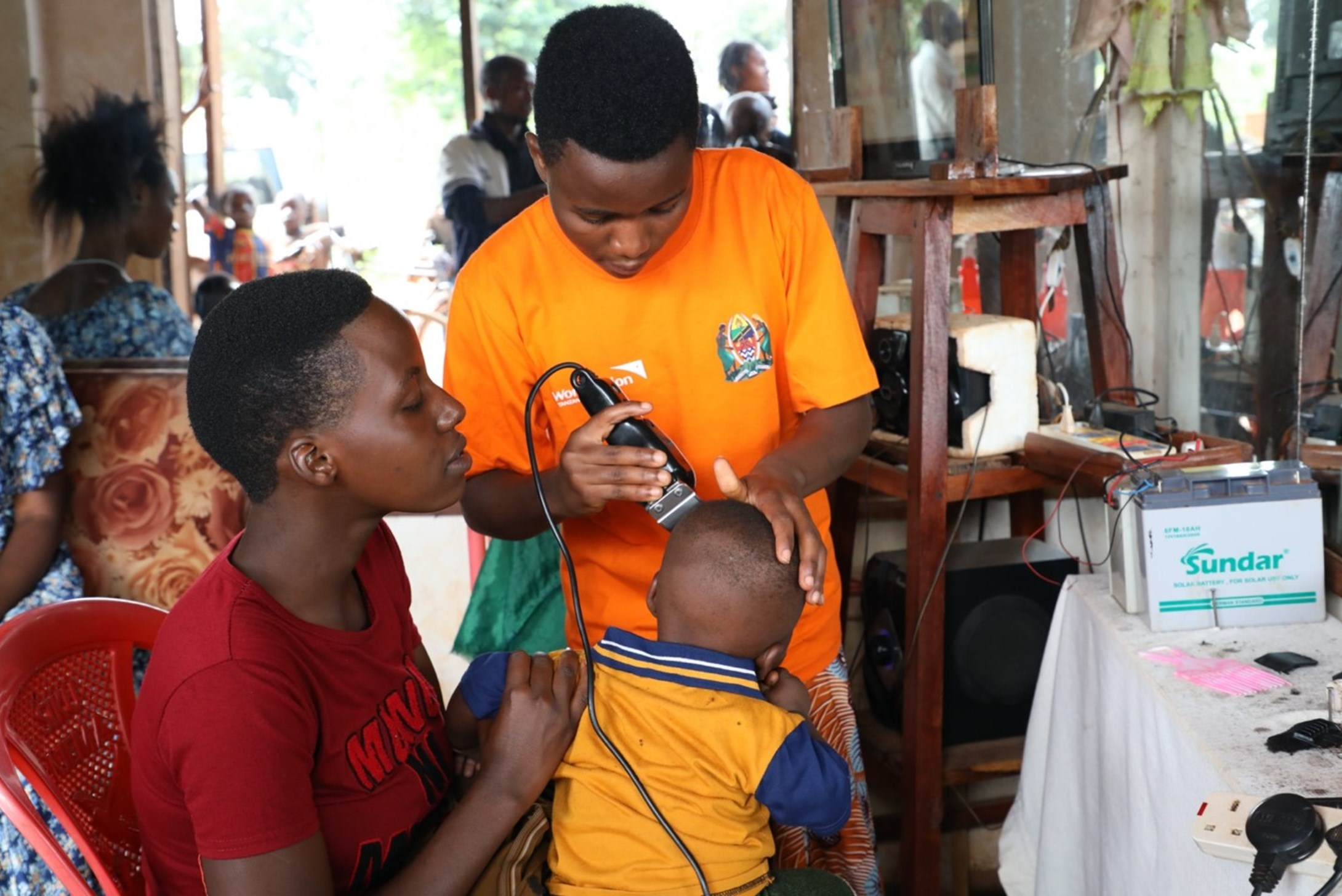 Girl in Tanzania shaving a child's head while his mother holds him.